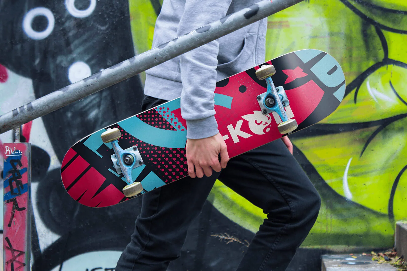 A skateboard carried past a graffiti wall, its deck printed in pink, teal and black with A-KON’s cat mascot