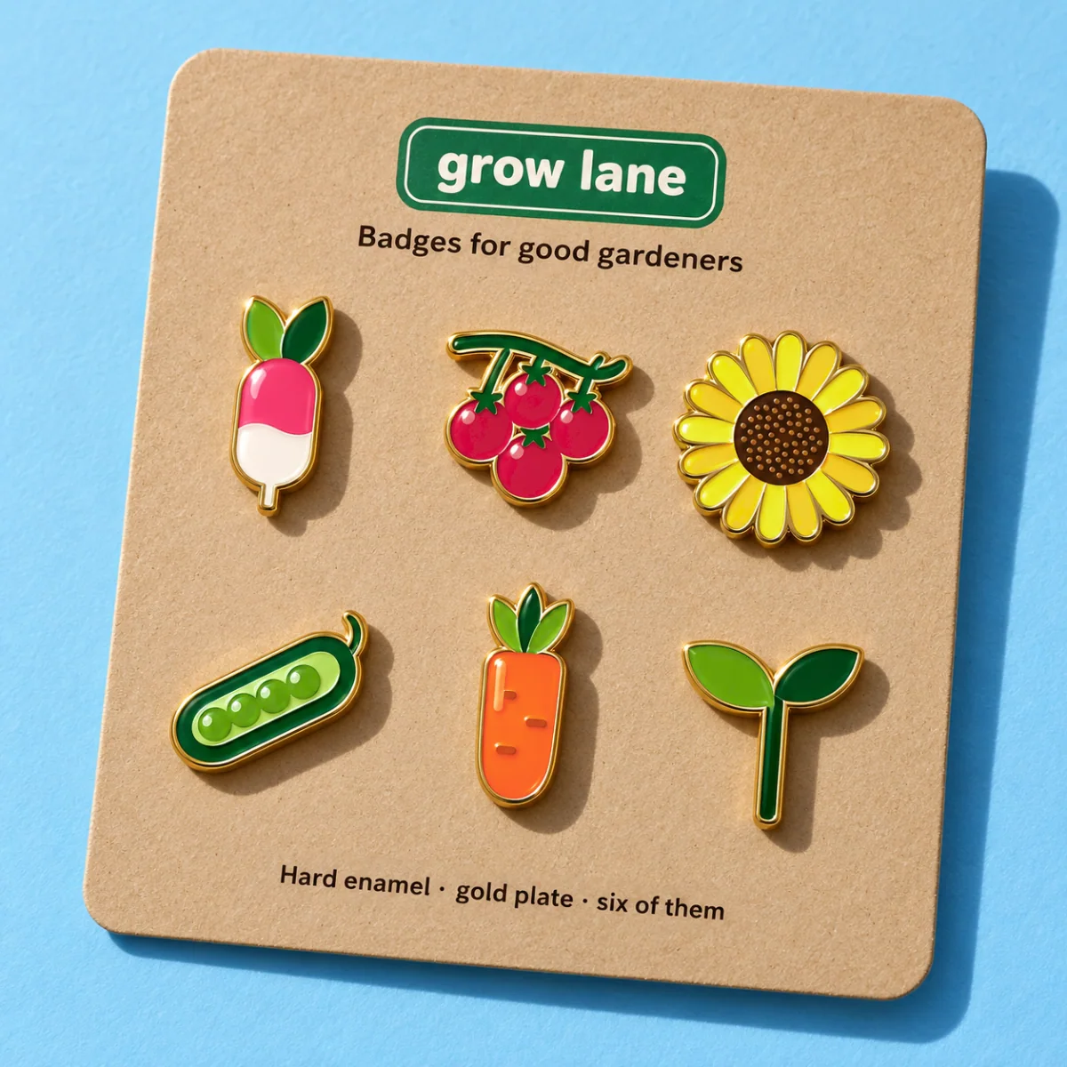 Six hard-enamel pins with gold edges on a kraft backing card on sky-blue paper: a radish, a truss of cherry tomatoes, a sunflower, a pea pod, a carrot and a seedling, under the green Grow Lane sign and “Badges for good gardeners”, in hard sun