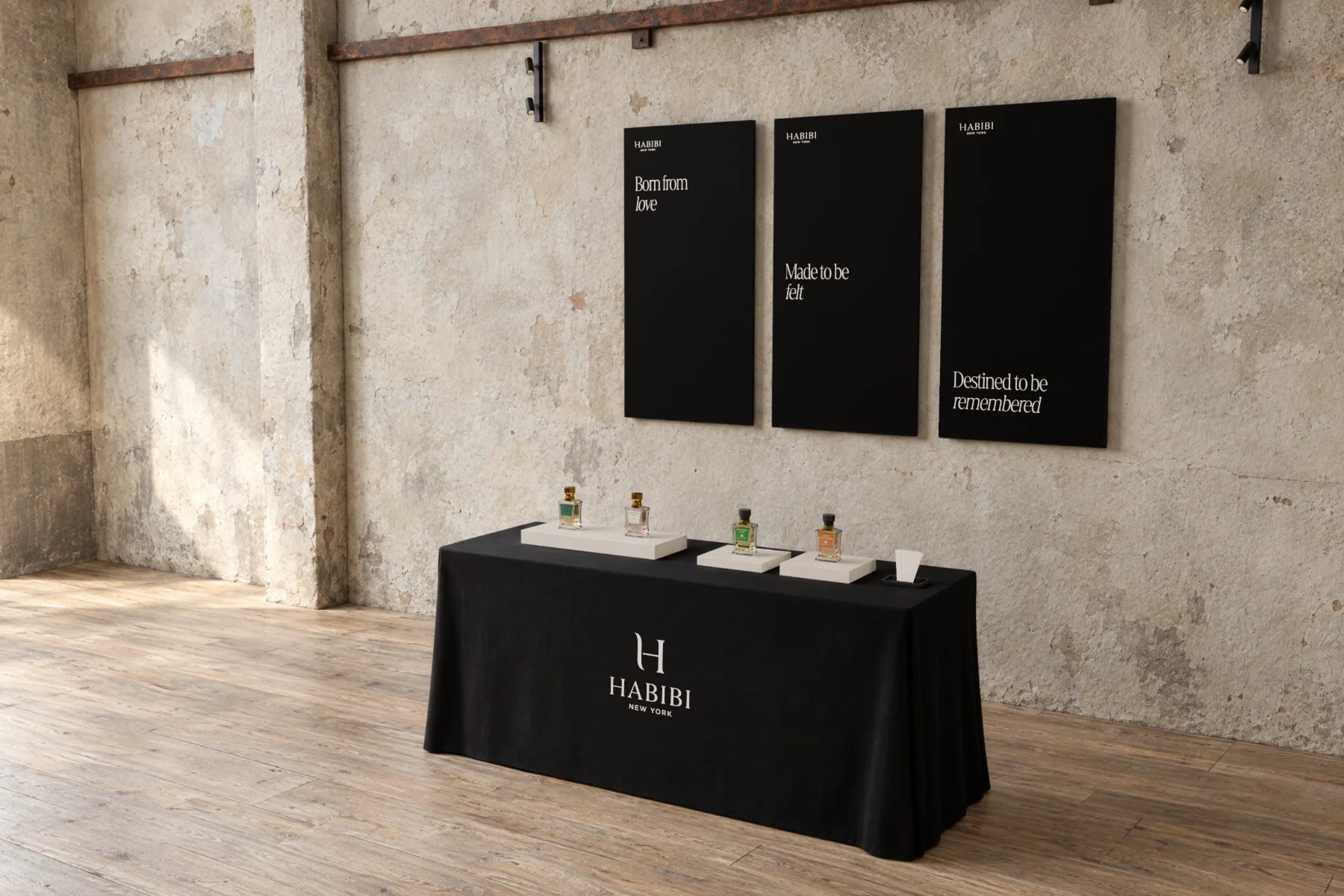 Habibi New York’s booth in a raw plaster hall: three black panels on the wall read “Born from love”, “Made to be felt” and “Destined to be remembered”, and below them a black-clothed table carries the H monogram and HABIBI NEW YORK, with the four extraits de parfum on white plinths