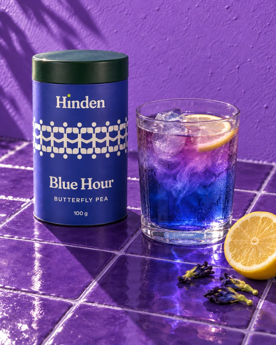 A tin of Hinden's Blue Hour tea, indigo blue with a cream band of the sprout mark repeated round it, standing on glossy violet tiles against a violet wall in hard sun, beside a glass of iced butterfly-pea tea, vivid blue at the bottom turning violet where a lemon wheel sits in it; a halved lemon and dried blue flowers on the tiles